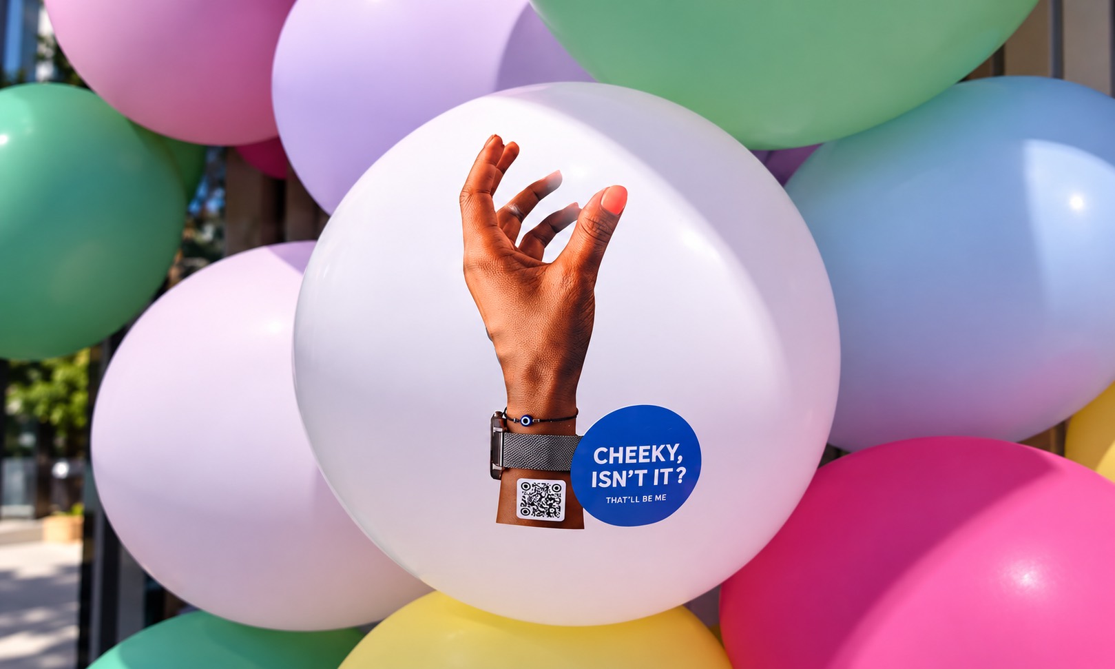A hand reaching up on a balloon among pastel balloons, with a 'Cheeky, isn't it?' badge